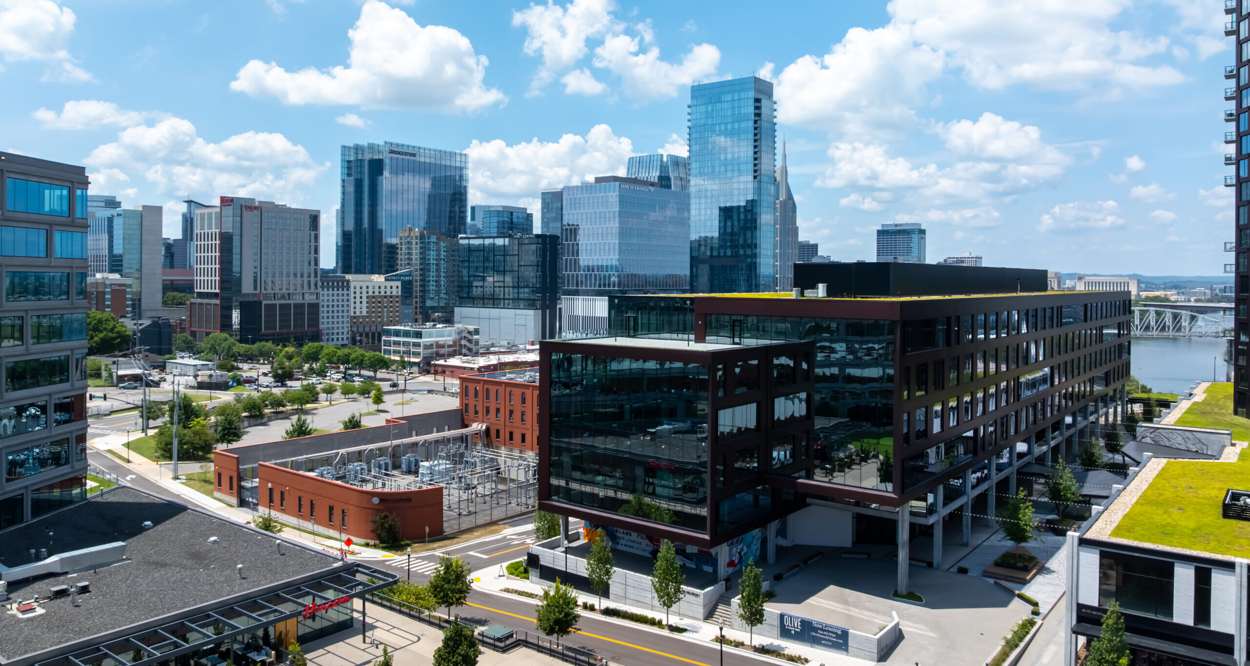 A cityscape with modern glass office buildings, a red brick building, green rooftops, and tree-lined streets under a blue sky with scattered clouds.