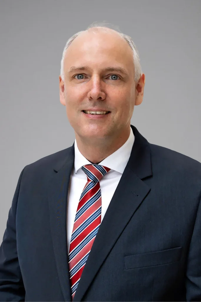 A man with short gray hair in a dark suit, white shirt, and red, navy, and white striped tie, smiling at the camera against a plain gray background.