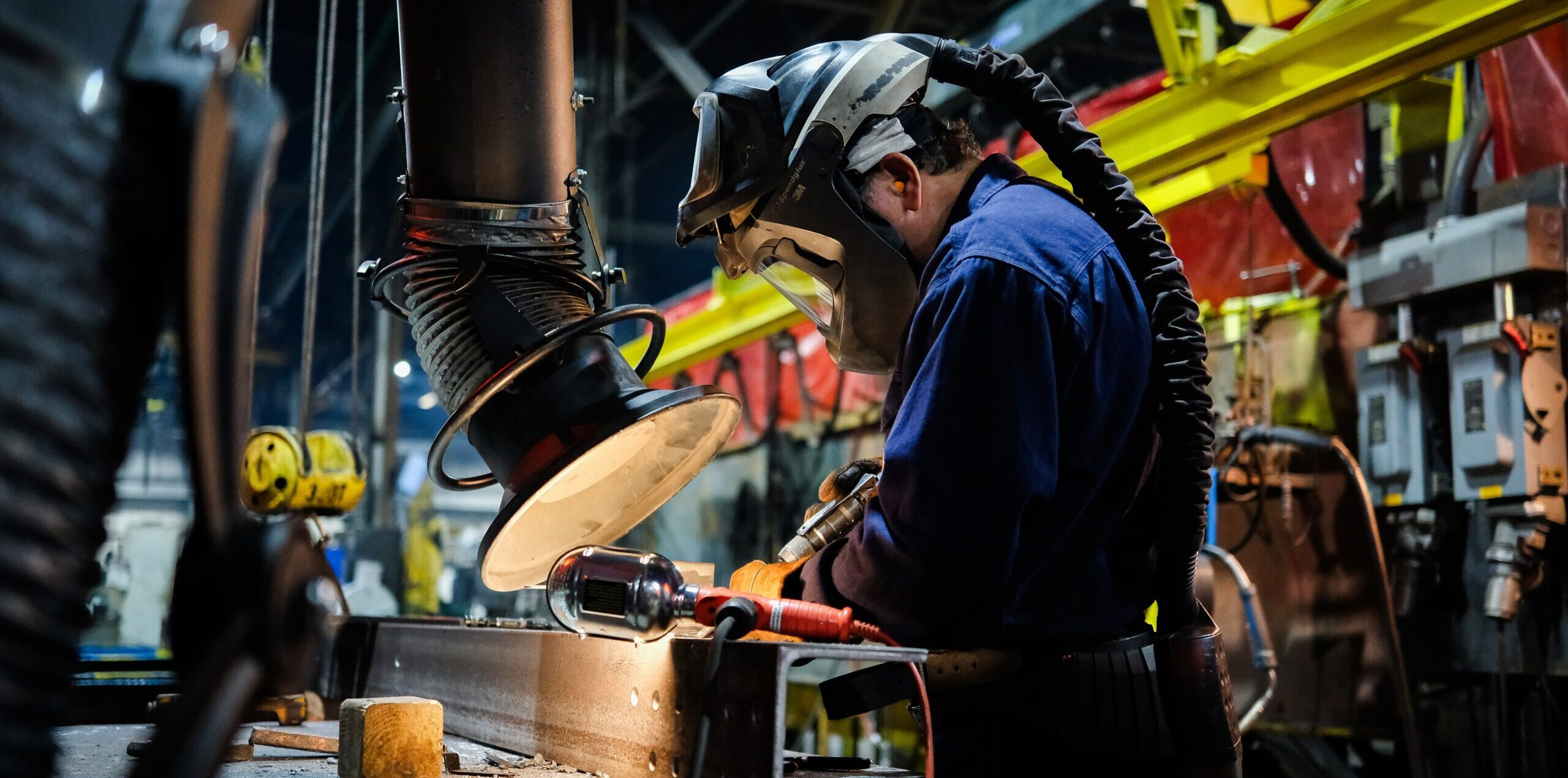 A worker wearing protective gear and a face shield uses tools to work on a metal beam in a factory setting, with industrial equipment and overhead lights visible in the background.