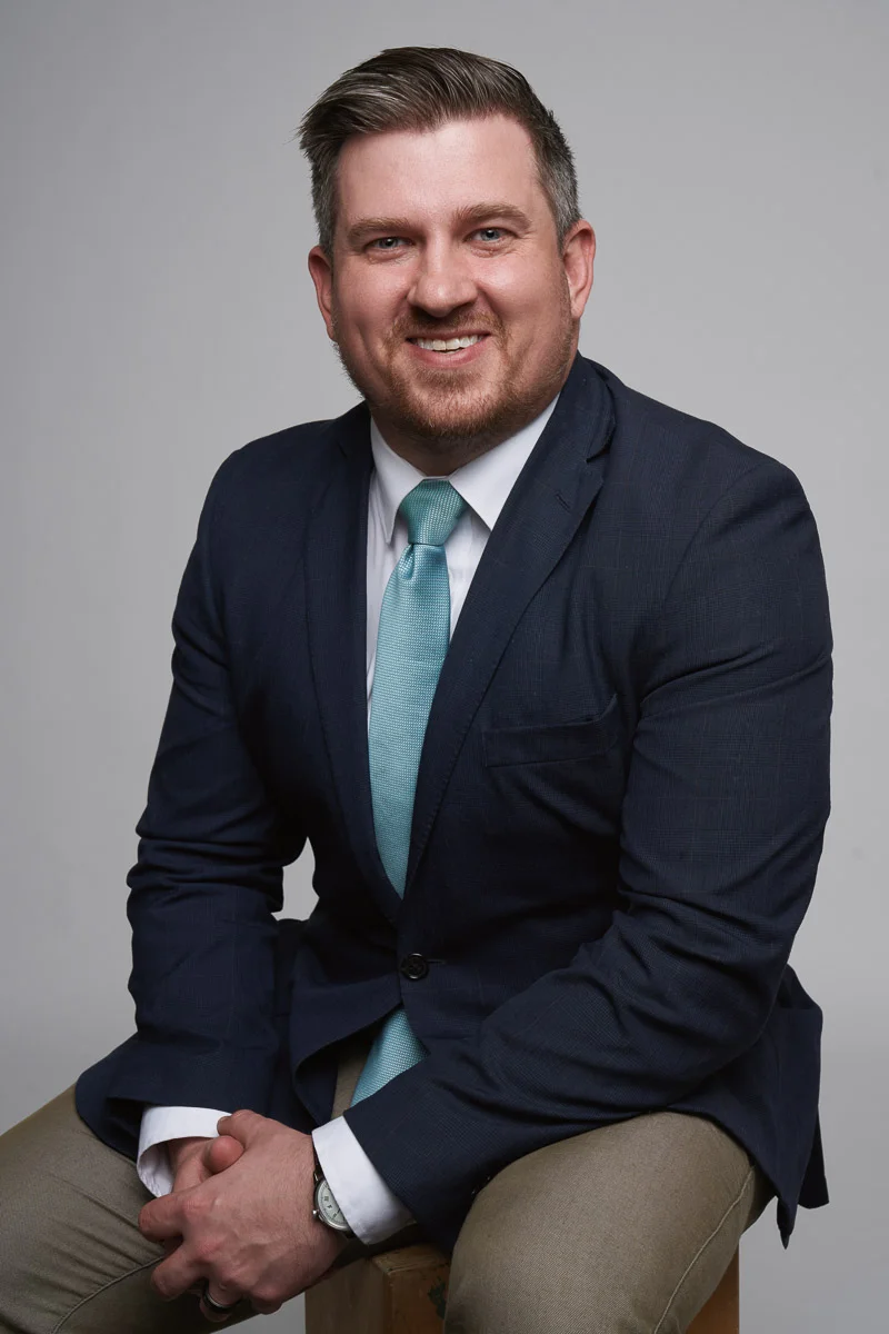 A man in a dark suit with a light blue tie sits on a stool, smiling at the camera against a plain gray background. He has short brown hair and a trimmed beard.