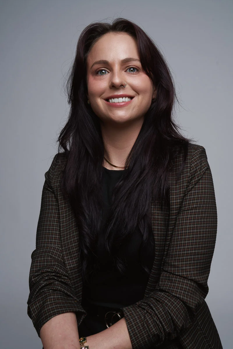 A woman with long dark brown hair wearing a checkered blazer and black top smiles at the camera against a plain gray background.