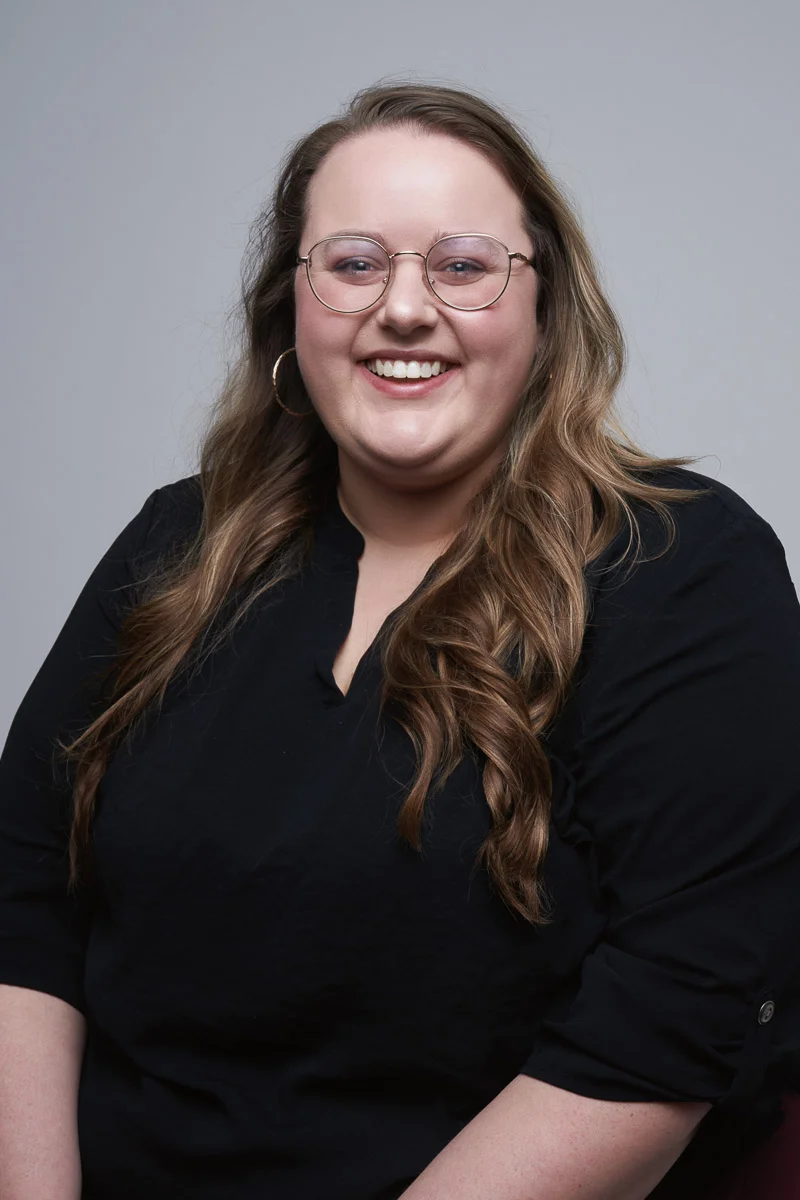 A smiling woman with long, wavy brown hair and glasses, wearing a black top, sits against a plain gray background.