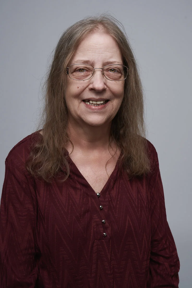 A woman with long brown hair and glasses smiles at the camera. She is wearing a maroon, patterned blouse and is posed against a plain, light gray background.