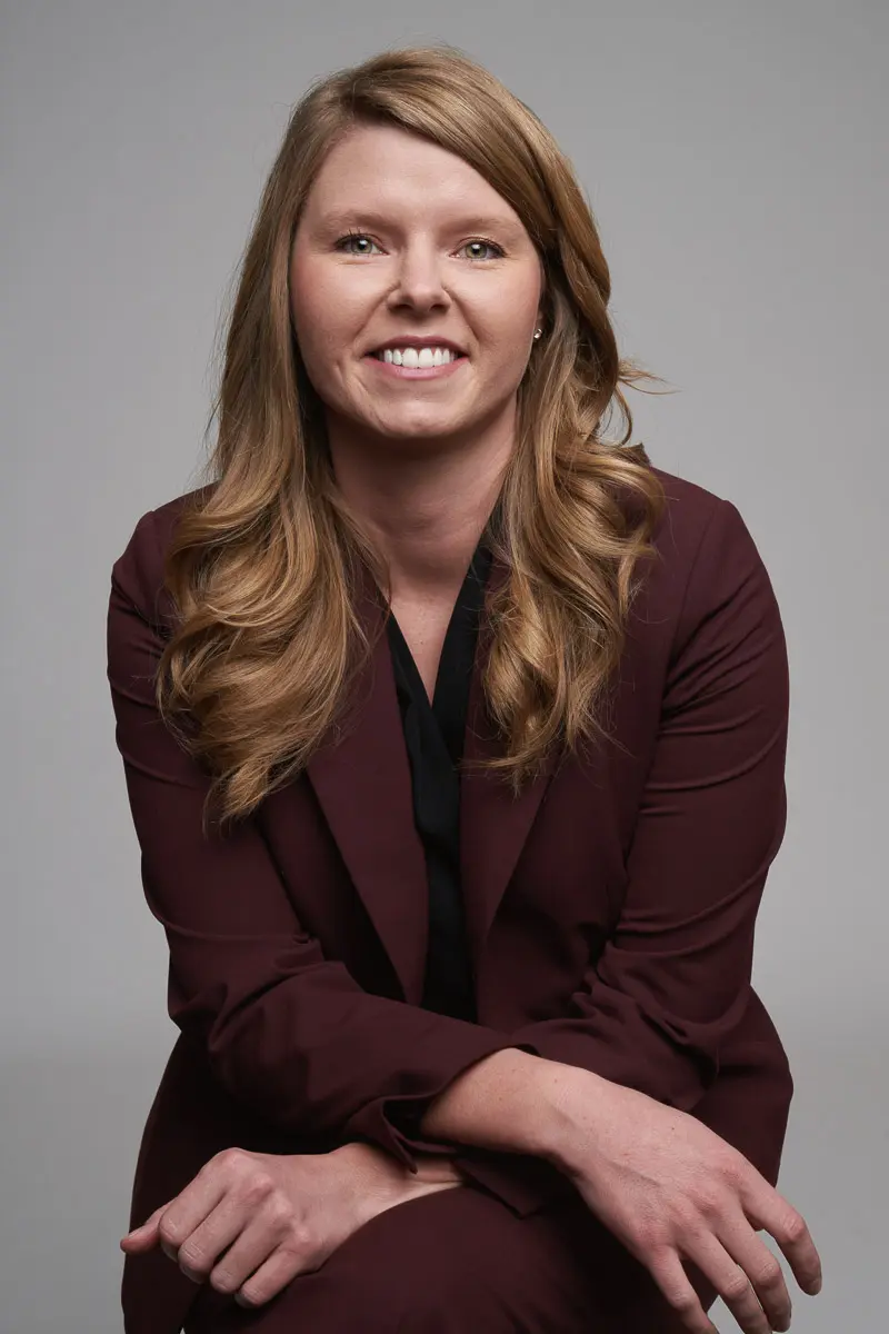 A woman with long, wavy blonde hair wearing a burgundy suit and black blouse sits on a stool, smiling confidently against a plain gray background.
