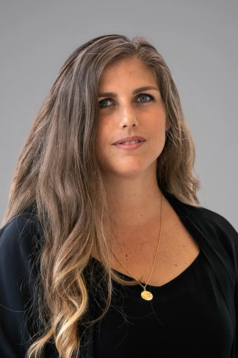 A woman with long, wavy brown hair wears a black top and a gold necklace with a pendant. She is looking at the camera with a neutral expression against a plain gray background.