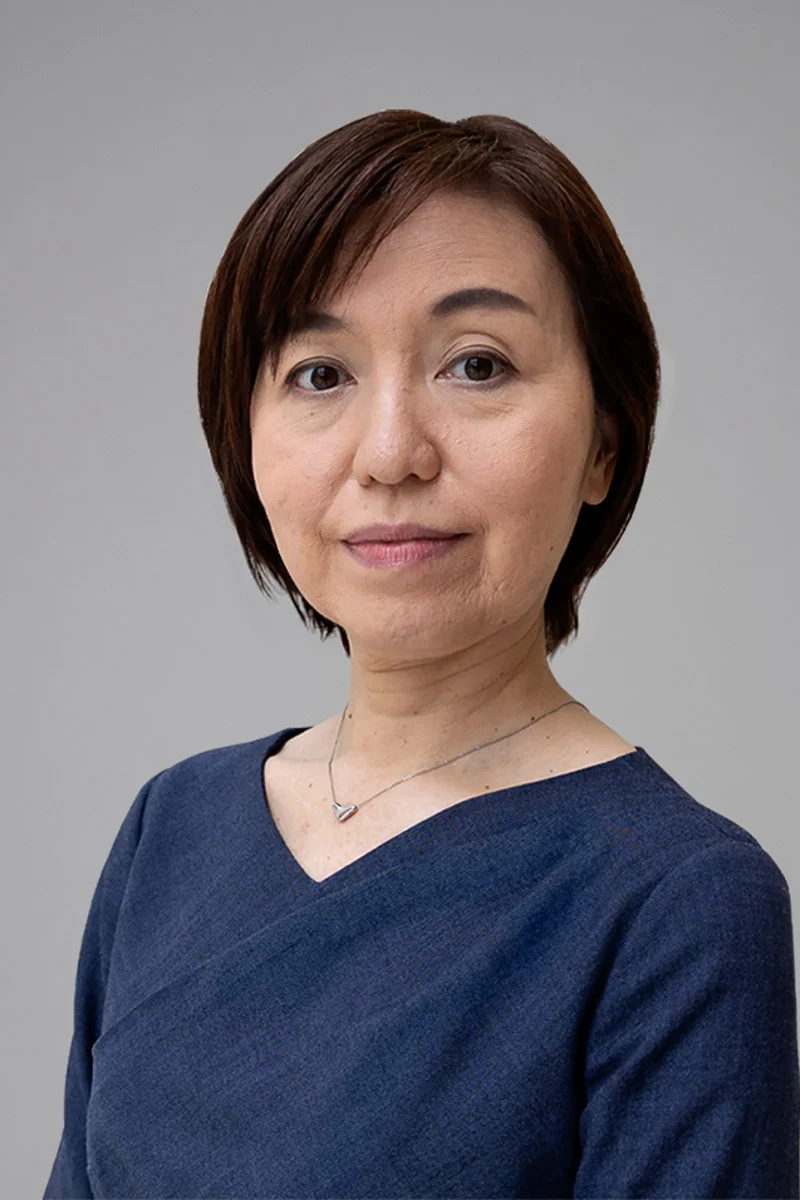 A woman with short brown hair, wearing a navy blue top and a silver necklace, stands against a plain light gray background, looking calmly at the camera.