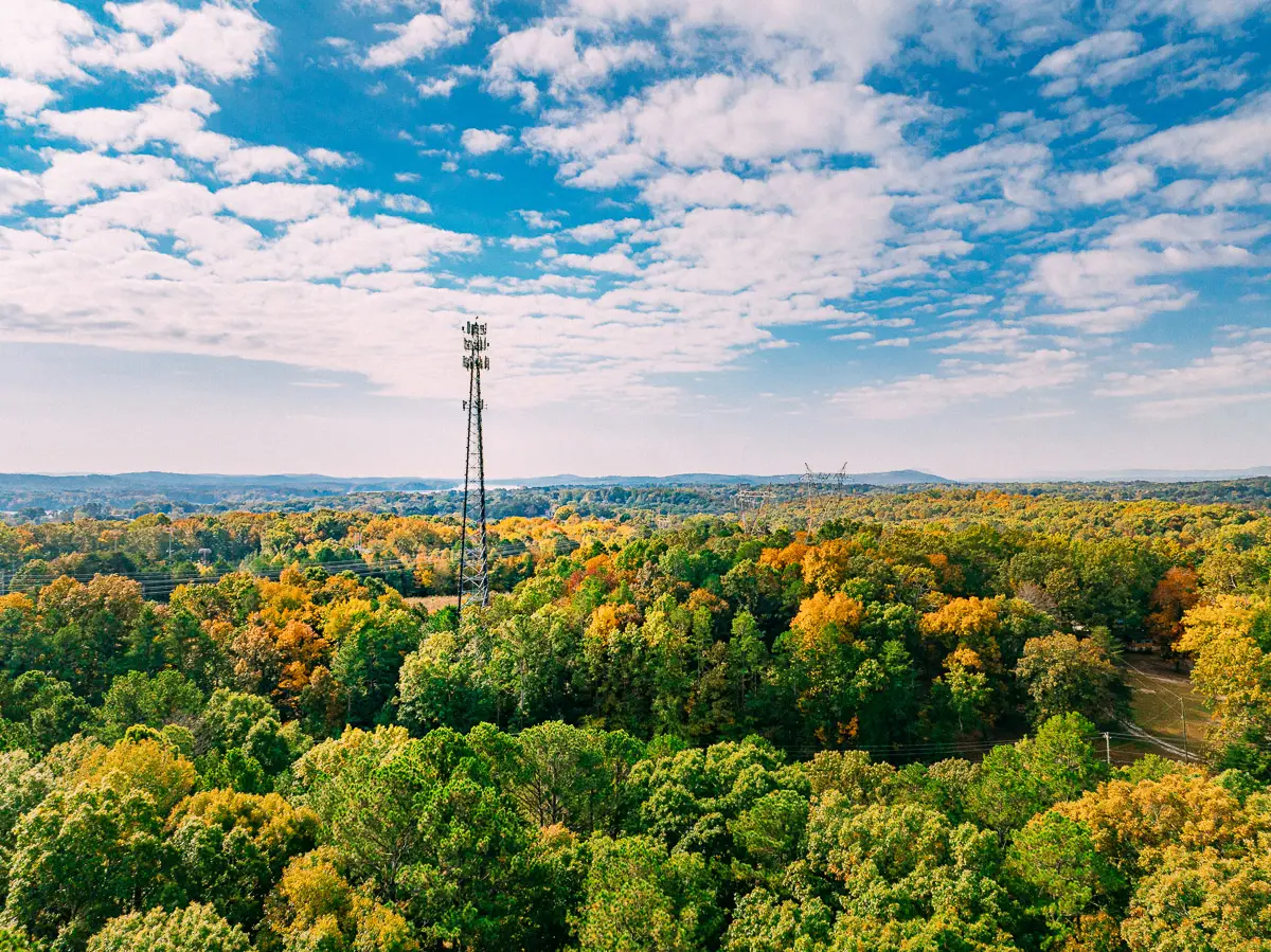 A tall cell tower rises above a dense forest with green and yellow autumn foliage under a partly cloudy blue sky, with distant hills visible on the horizon.