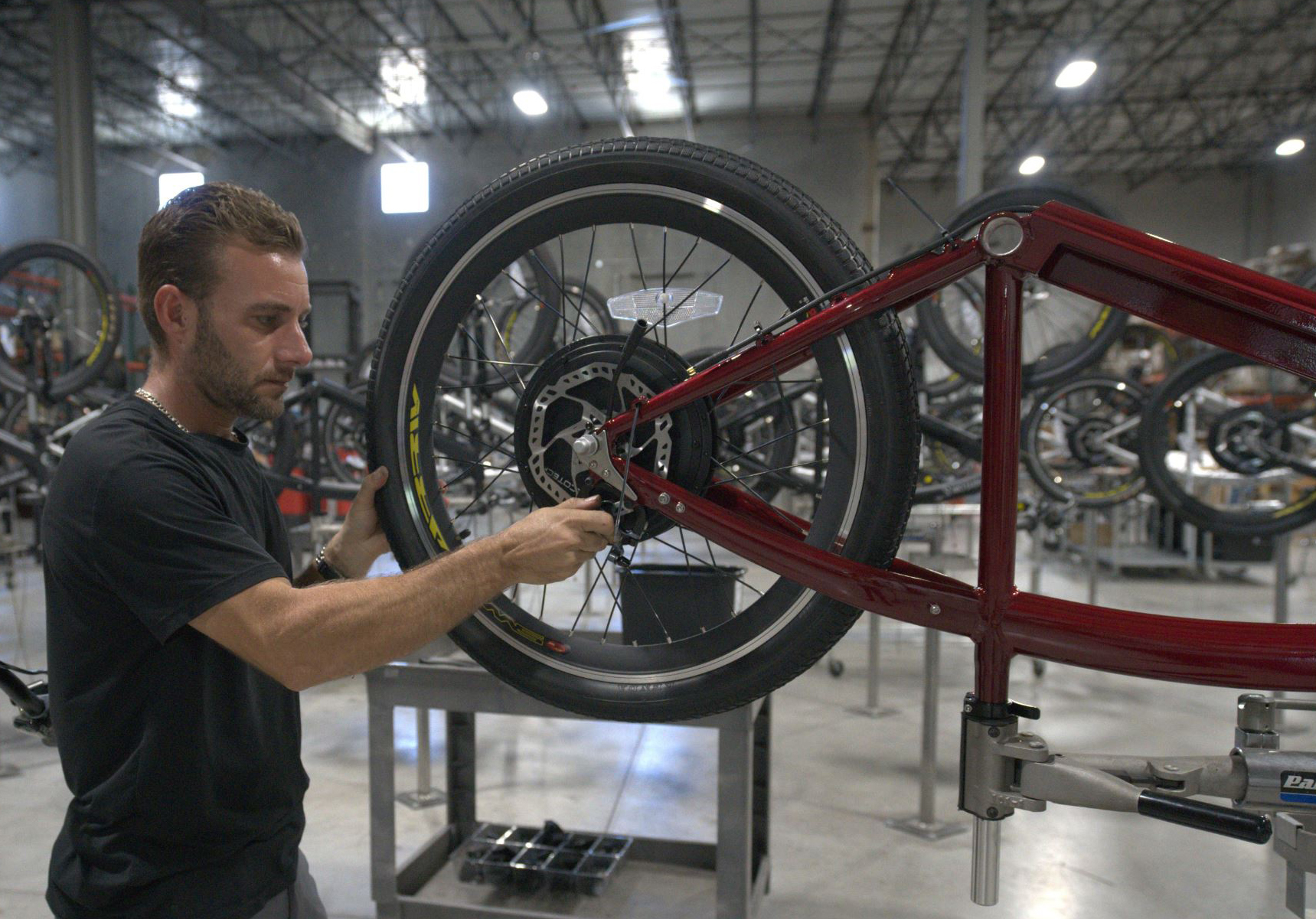 A man in a workshop is assembling a red bicycle, adjusting the rear wheel. The background shows rows of bicycles and industrial lighting in a large, spacious facility.