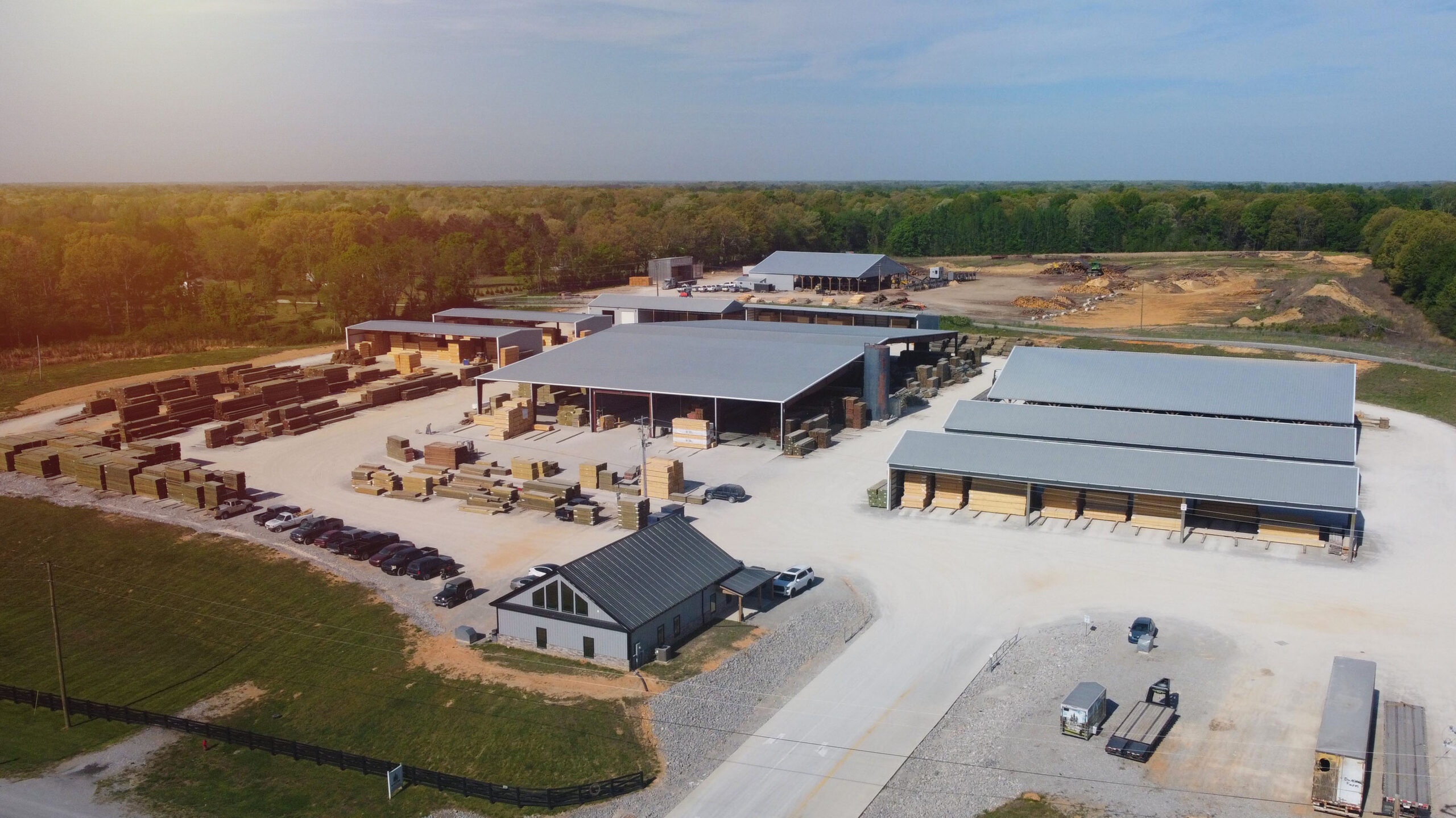 Aerial view of a large lumberyard with stacks of wood, multiple metal-roofed buildings, parked cars, trucks, and surrounding green forest under a clear sky.