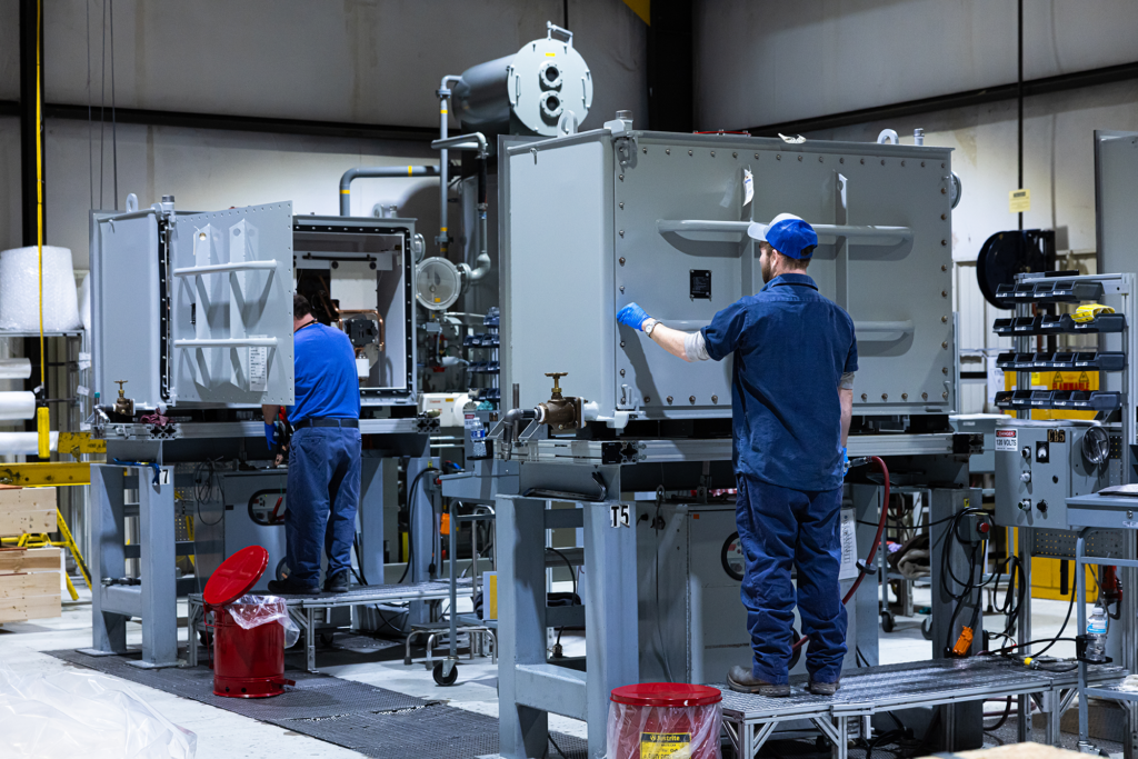 Two workers in blue uniforms and caps operate large industrial machines in a well-lit factory setting, surrounded by equipment and machinery.