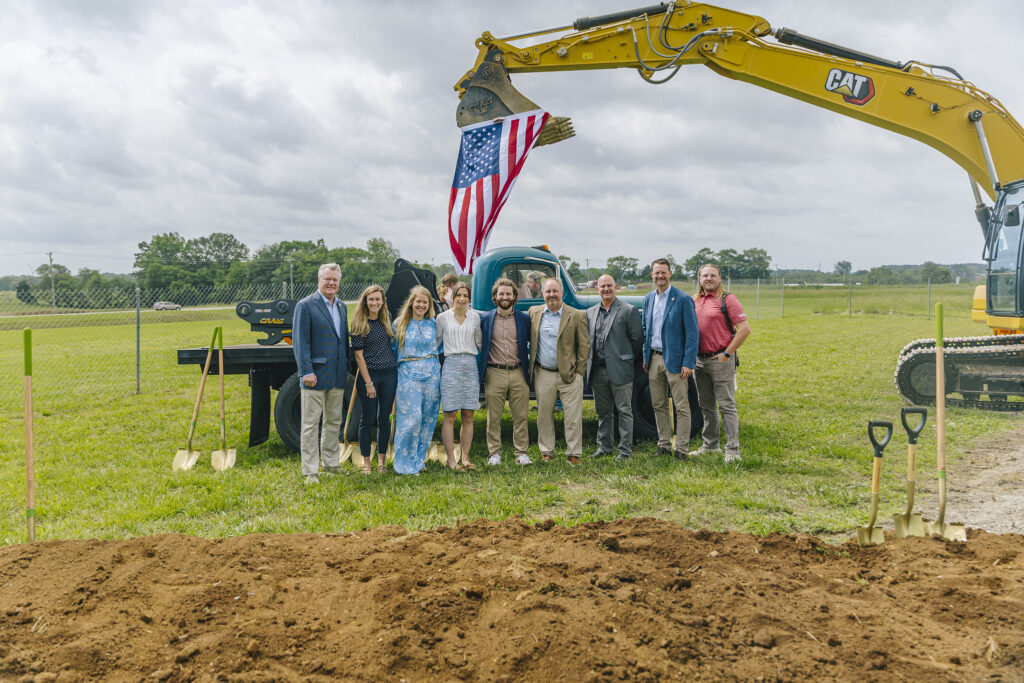 A group of nine people in business attire stand in front of a large yellow excavator with an American flag hanging from its arm, on a grassy field next to a row of shovels stuck in the dirt.