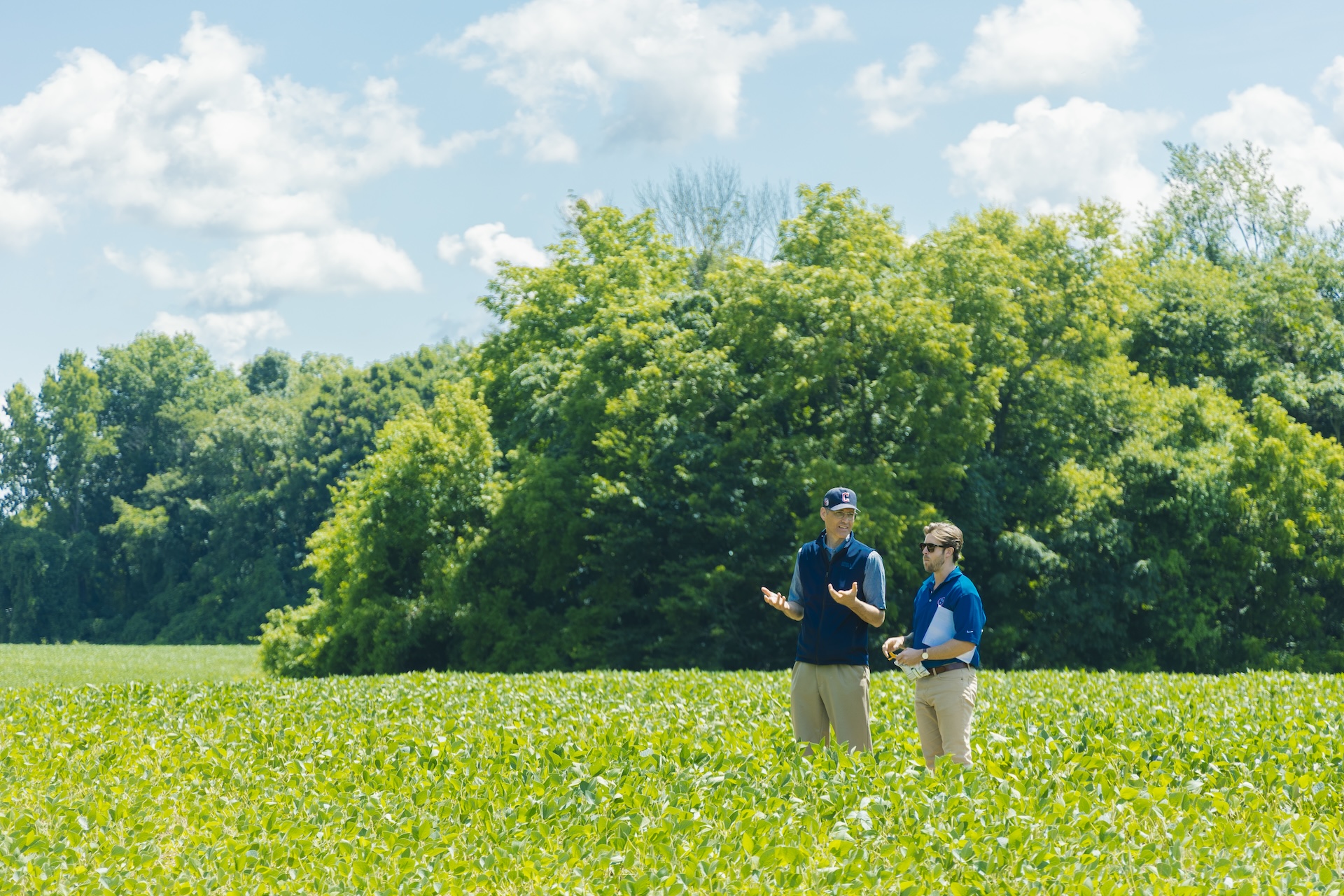 Two people in blue shirts and khaki pants stand in a green crop field, talking. Lush trees and a bright blue sky with clouds are in the background.