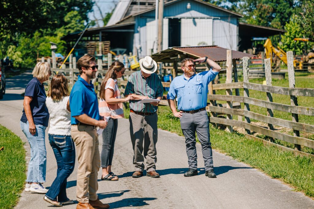 A group of six adults stands on a rural road by a wooden fence; some are looking at papers or maps, while others chat. A barn and green trees are visible in the background on a sunny day.