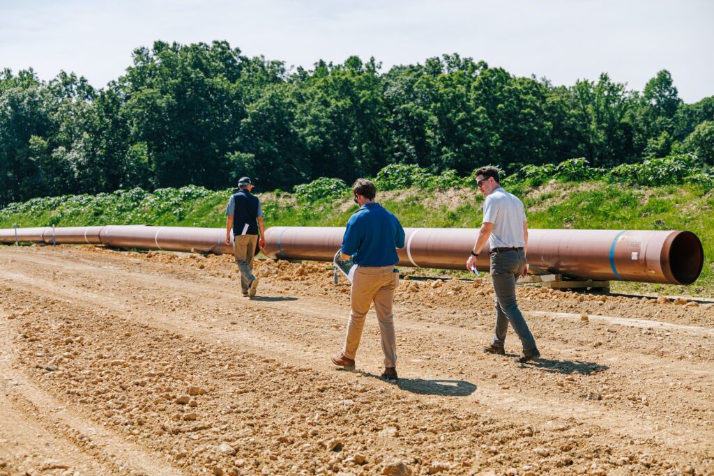Three men in business attire walk along a dirt road next to a large metal pipeline under construction, with green trees and plants in the background.