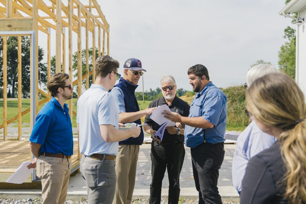 A group of people stand outdoors near a wooden building frame, reviewing papers and discussing plans on a sunny day. Some wear sunglasses and blue shirts, and trees are visible in the background.