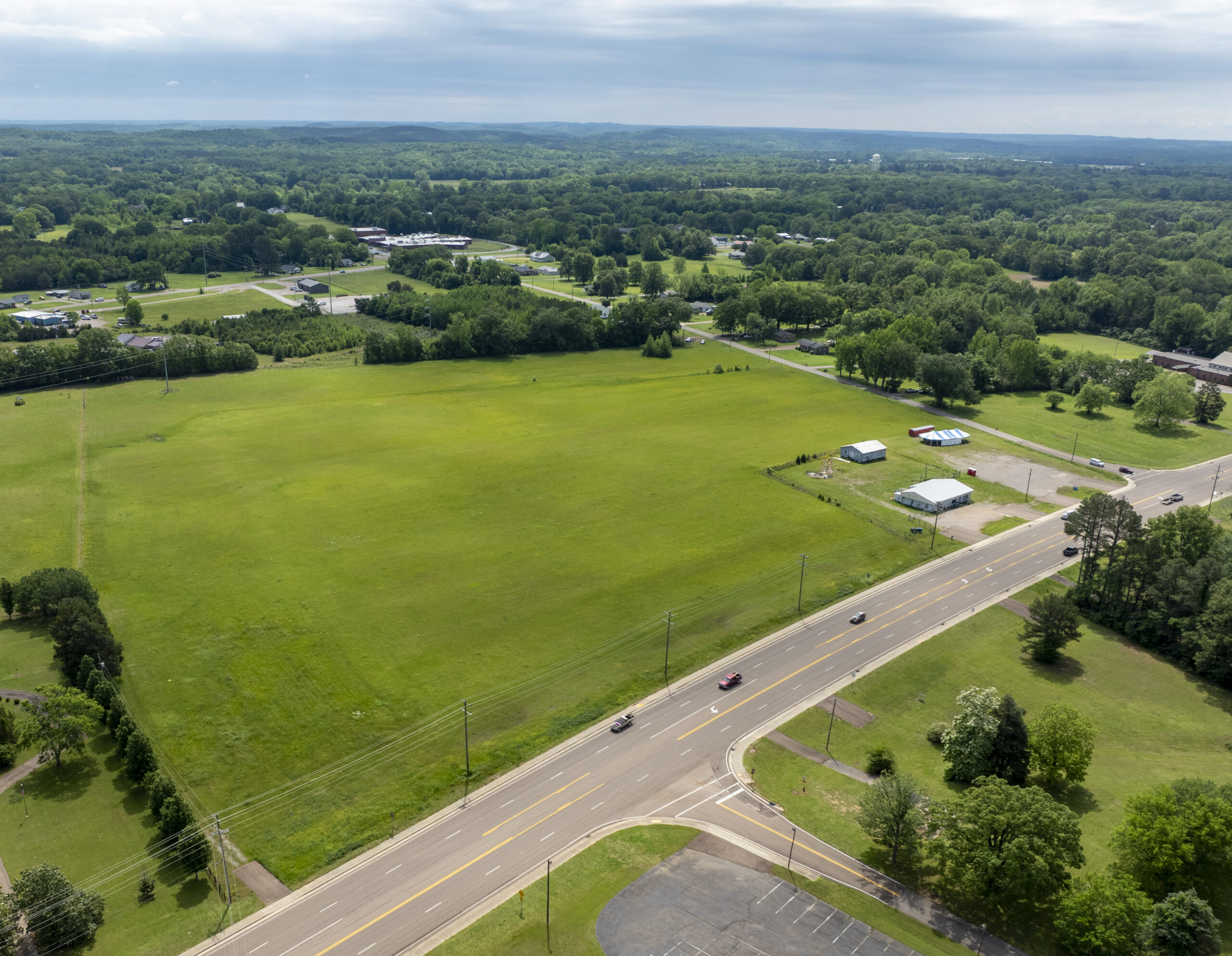 Aerial view of a wide, open green field bordered by a road with cars, scattered trees, small buildings, and distant forested hills under a cloudy sky.