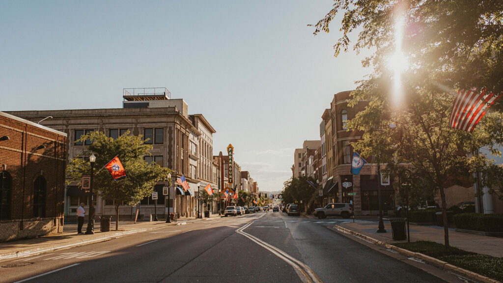 A sunlit downtown street with historic buildings, cars parked along the sides, and various colorful flags hanging from lampposts. The sunlight filters through trees, and an American flag is visible on the right.