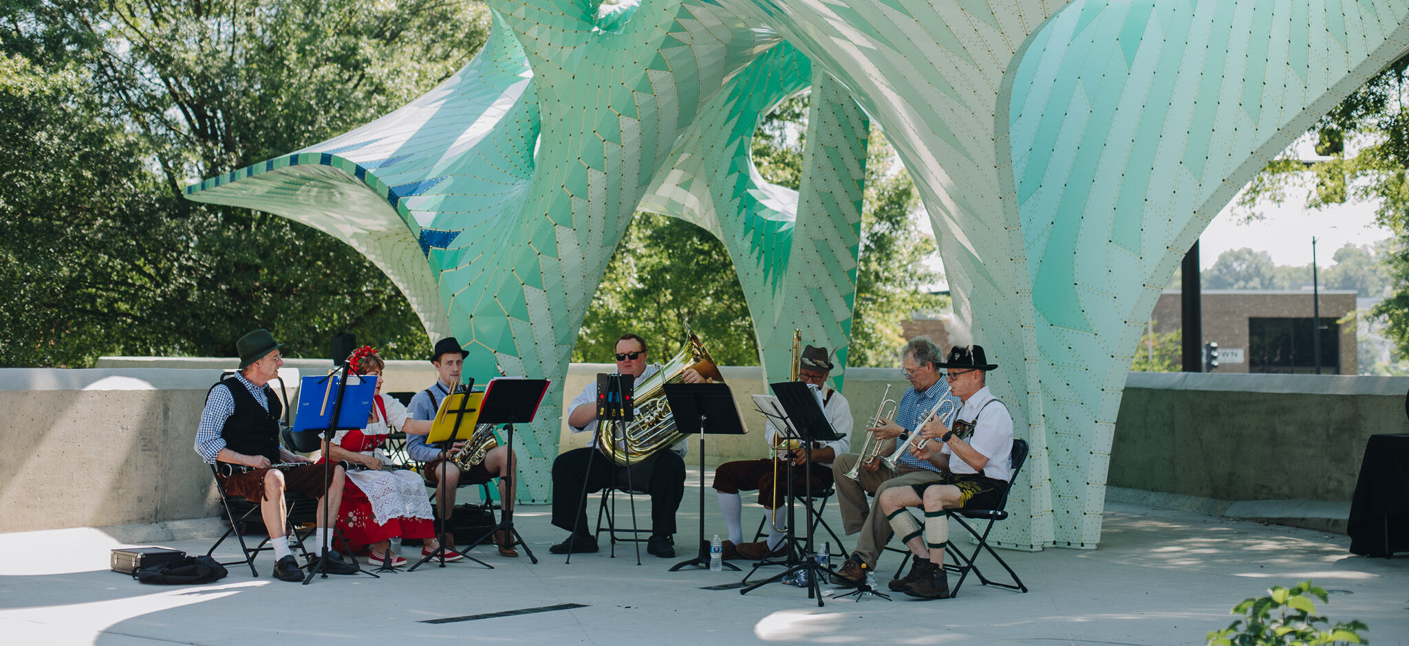 A small band in traditional Bavarian attire performs outdoors under a large, modern, turquoise and white sculptural canopy, with trees and sunlight in the background.