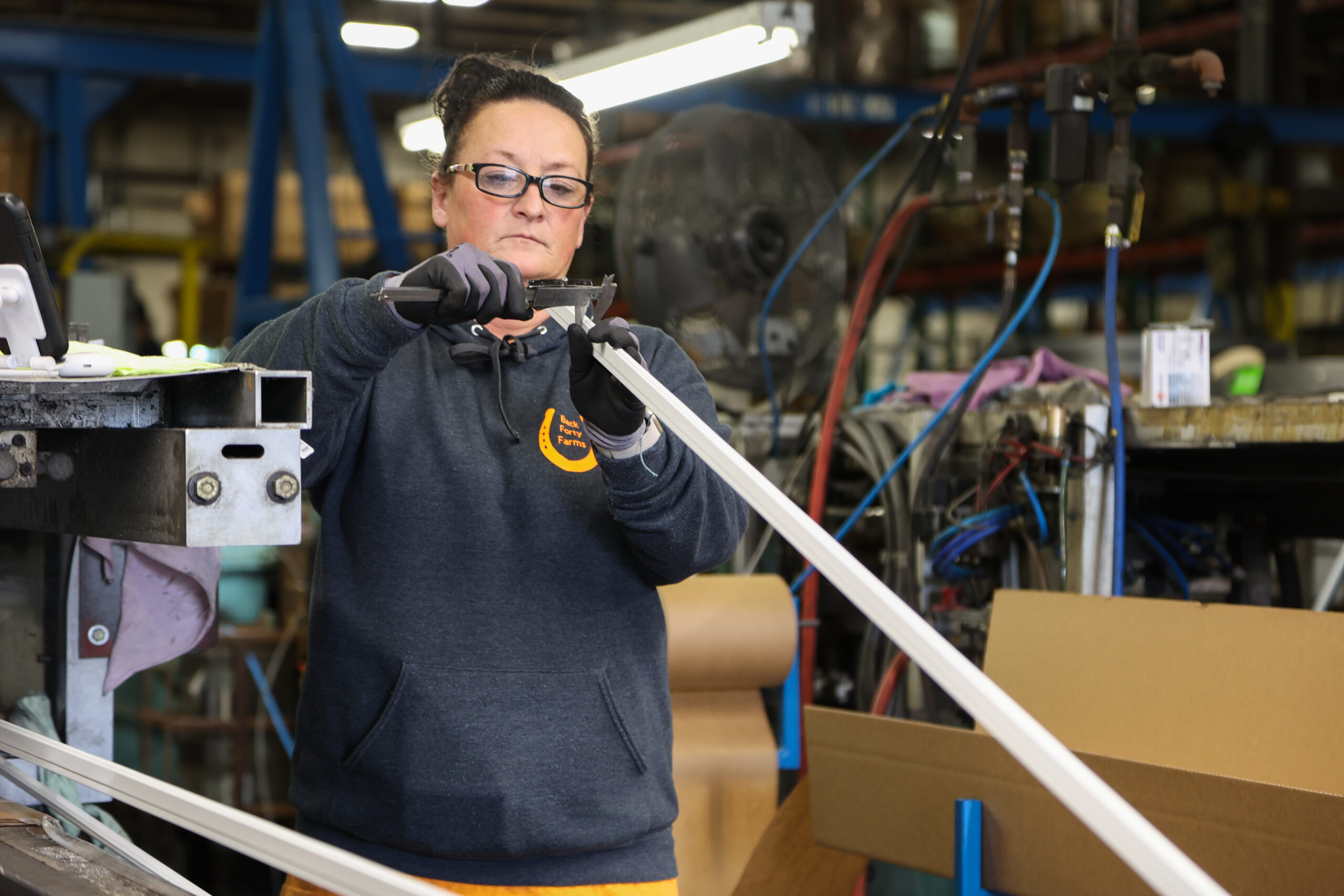 A woman wearing glasses and gloves works in a factory, using a tool to measure or trim a long metal or plastic component, surrounded by industrial equipment and boxes.