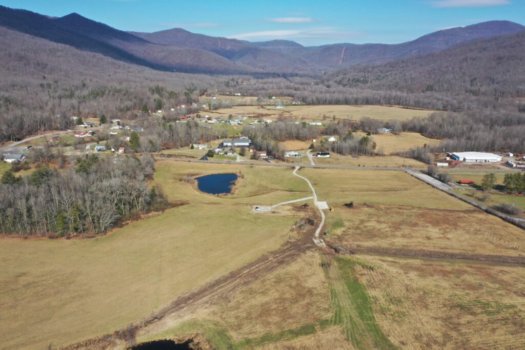 Aerial view of a rural valley with scattered houses, ponds, open fields, and surrounding forested hills under a clear blue sky. Roads and patches of trees cross the landscape.