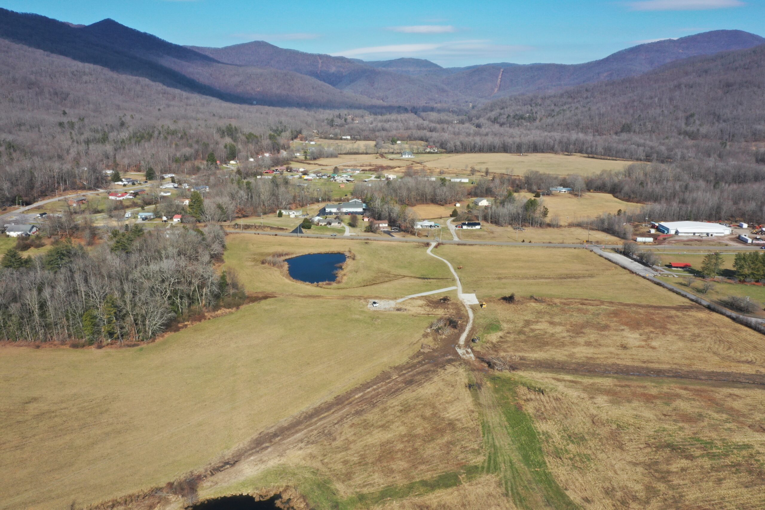 Aerial view of a rural valley with scattered houses, ponds, open fields, and surrounding forested hills under a clear blue sky. Roads and patches of trees cross the landscape.