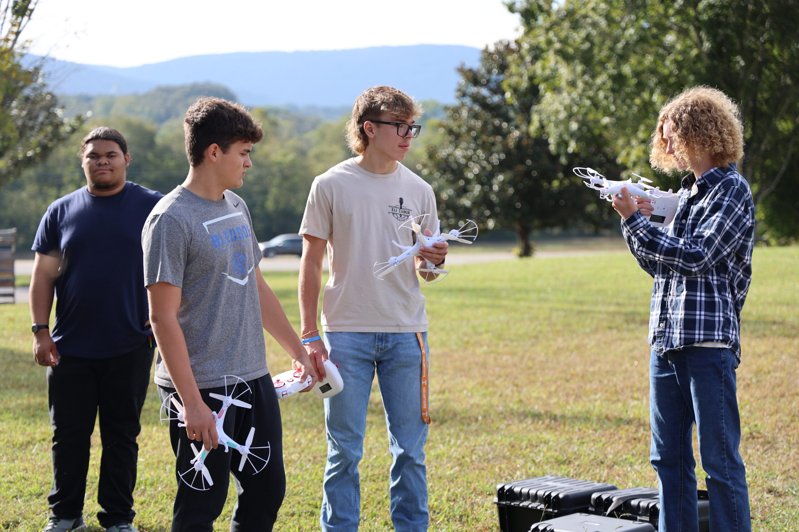 Four teenagers stand on a grassy field, holding drones and remote controls, talking and preparing for flight. Trees and mountains are visible in the background on a sunny day.