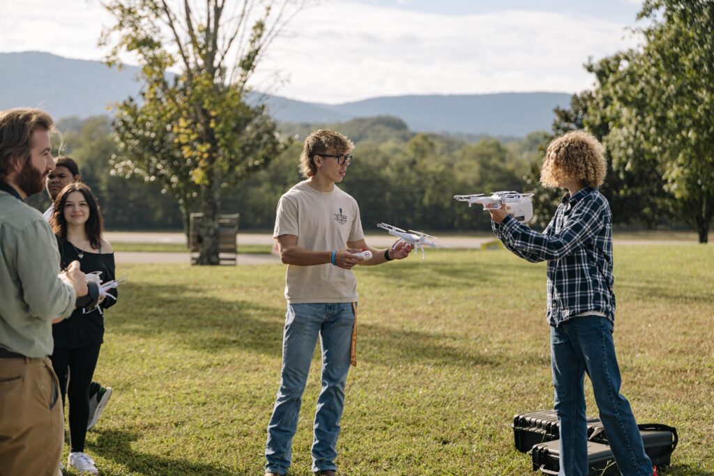 A group of people standing on grass, two of them holding drones and talking, with open cases nearby. Trees and mountains are visible in the background under a partly cloudy sky.