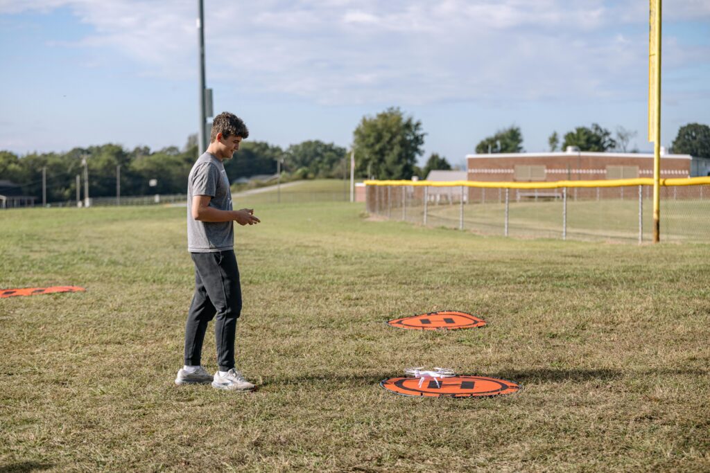 A person operates a small white drone on a grassy field, standing near orange landing pads. There is a fence, a yellow pole, and a building in the background under a partly cloudy sky.