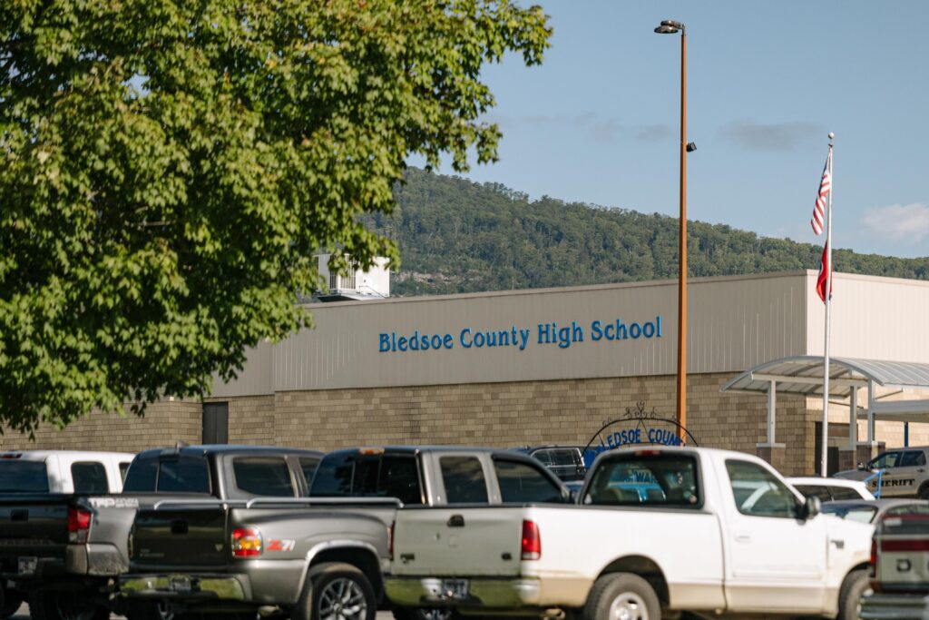Several pickup trucks are parked in front of Bledsoe County High School, a light-brick building with blue lettering. An American flag and a tree are also visible in the foreground.