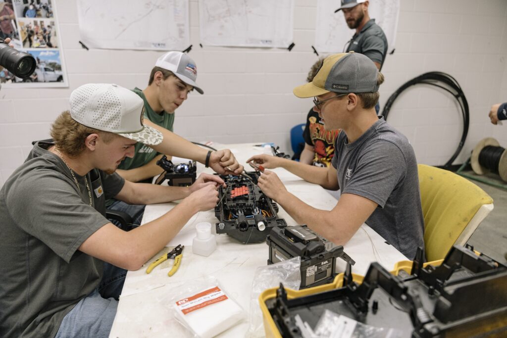 Three young men in baseball caps work together assembling or repairing electronic equipment at a table covered with tools and parts in a workshop. Another person stands in the background.