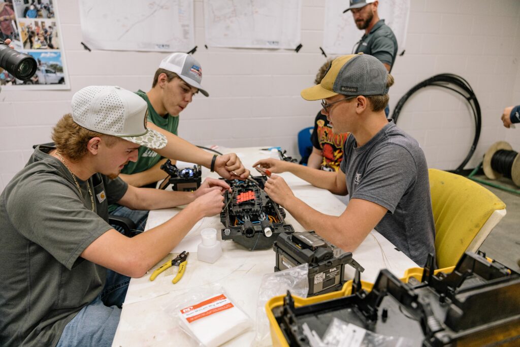 Three young men wearing caps work together on assembling or repairing an electronic device at a table, surrounded by tools and components, in a workshop or classroom setting.