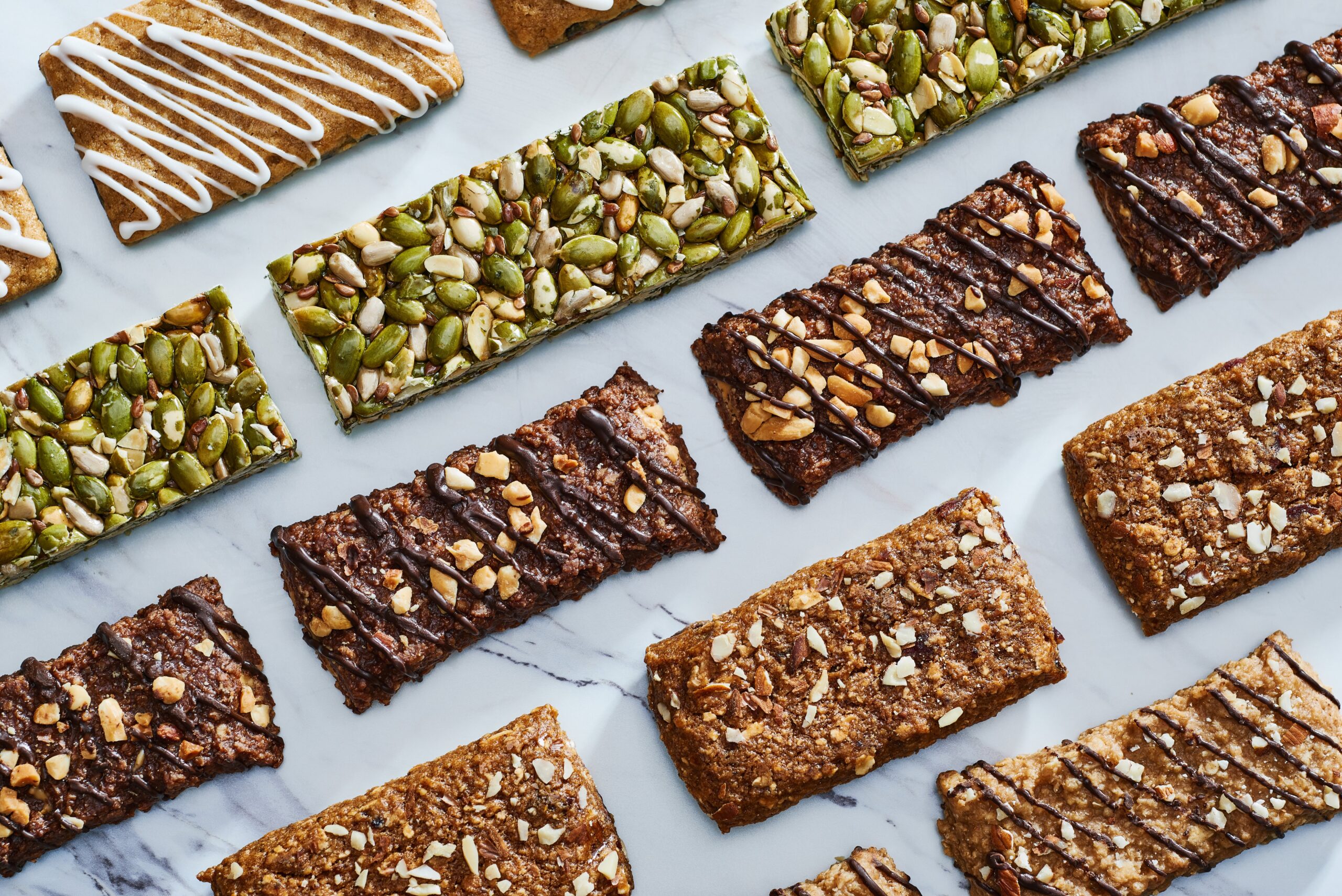 A variety of granola and protein bars with nuts, seeds, and icing, arranged in rows on a marble surface.