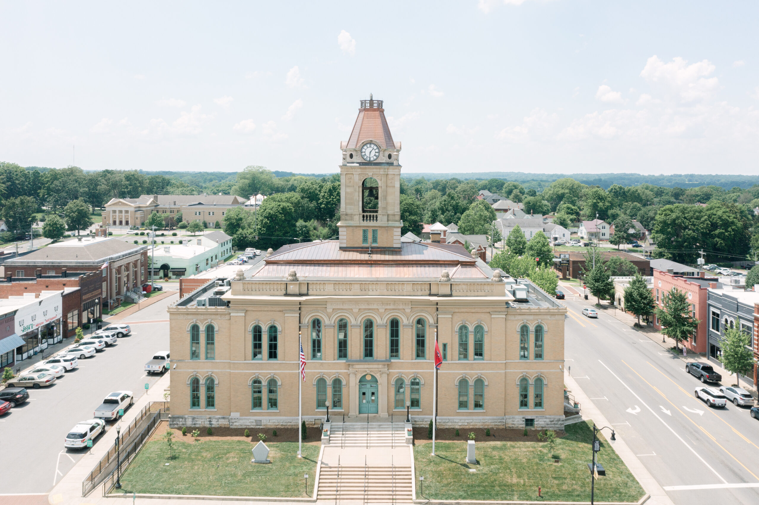 Aerial view of a historic courthouse with a clock tower, surrounded by parked cars, trees, and nearby buildings on a sunny day.