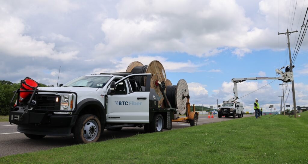 A BTC Fiber truck with large cable spools is parked by a road as workers install or repair lines on utility poles using a bucket truck. The scene is under a partly cloudy sky.