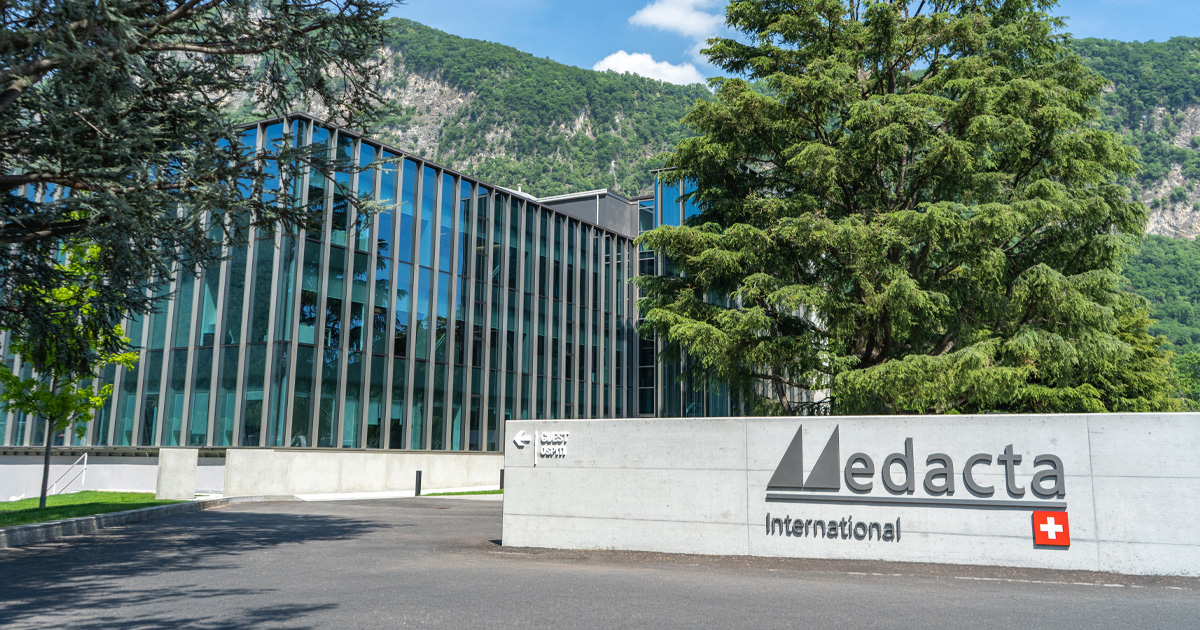 Modern glass office building with a large tree in front and a sign reading “Medacta International” with a Swiss flag, set against green hills and a blue sky.