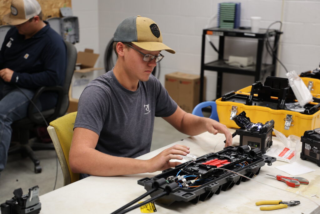 A person wearing glasses and a baseball cap works on assembling or repairing electronic equipment at a table in a workshop, surrounded by tools and components.