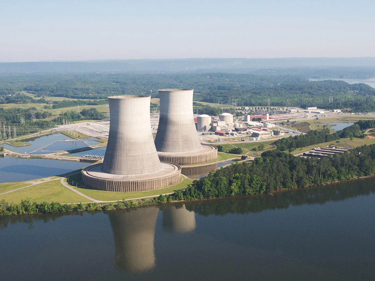 Aerial view of a nuclear power plant with two large cooling towers near a river, surrounded by greenery and additional buildings in the background.