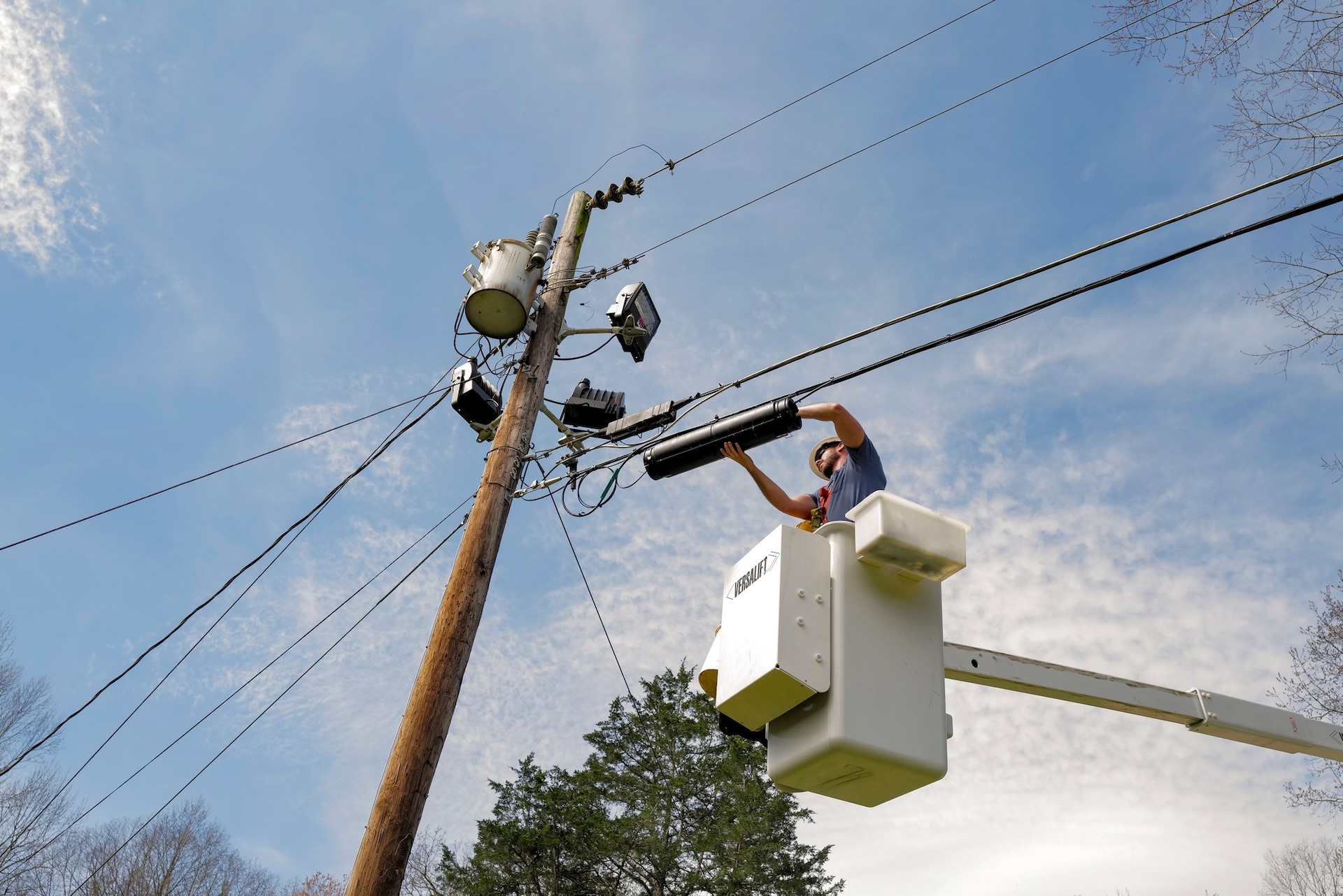 A worker in a bucket lift repairs power lines on a tall wooden utility pole against a blue sky with thin clouds and tree branches visible in the background.
