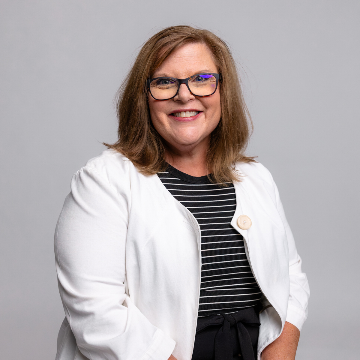 A woman with shoulder-length brown hair and glasses, wearing a white blazer over a black and white striped top, smiles at the camera against a plain light gray background.