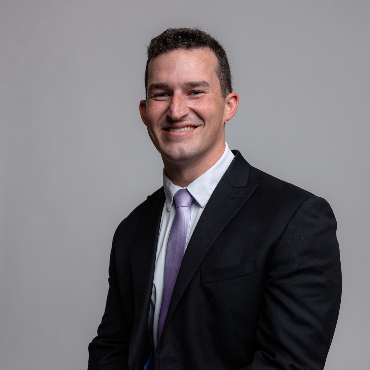 A man in a black suit, white shirt, and light purple tie is smiling while posing against a plain gray background.