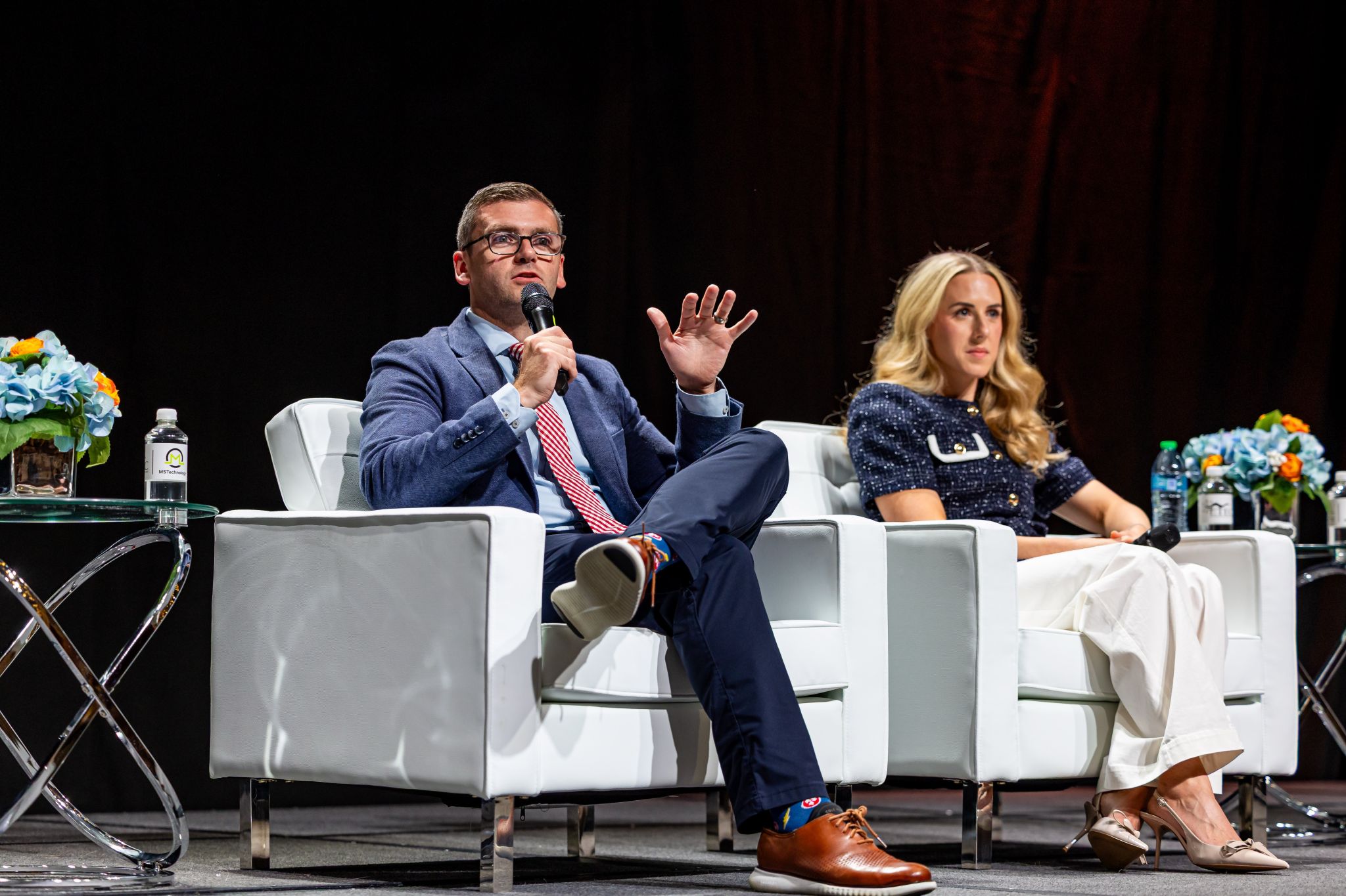 A man in a blue suit speaks into a microphone while seated on stage next to a woman in a navy top and white pants. Both sit in white chairs with flower arrangements on tables nearby.