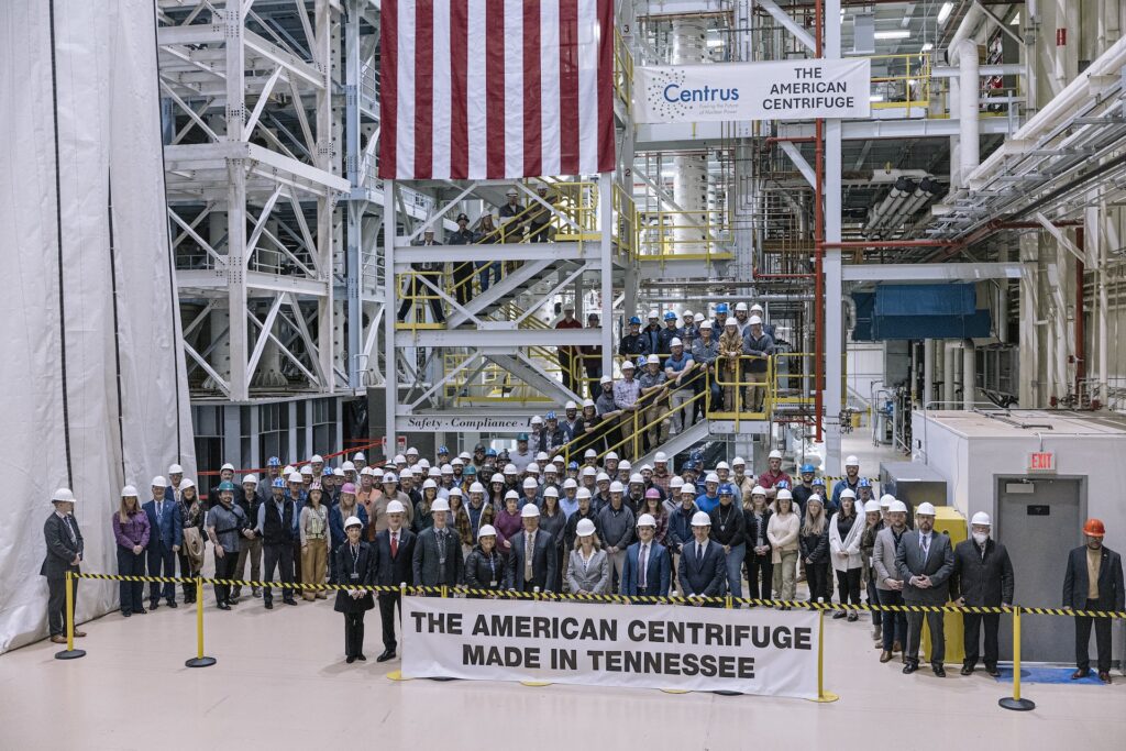 A large group of people in hard hats stand behind a sign reading “THE AMERICAN CENTRIFUGE MADE IN TENNESSEE” in an industrial facility, with safety barriers, metal stairs, and a U.S. flag in the background.