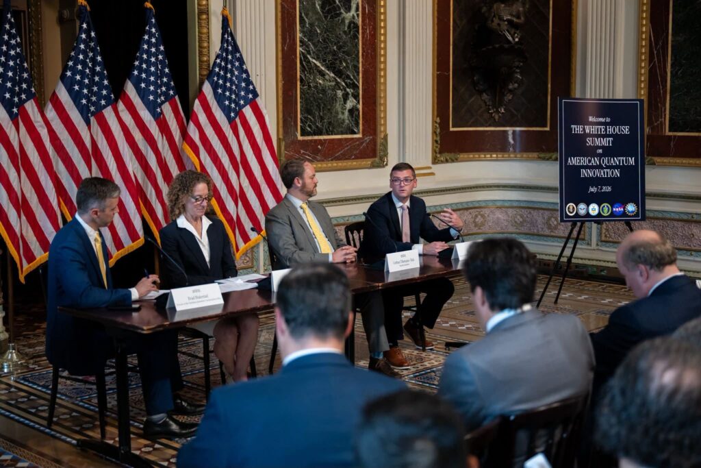 Four panelists sit at a table discussing at the White House Summit on American Quantum Innovation, with U.S. flags and a signboard in the background; audience members are seated in the foreground.
