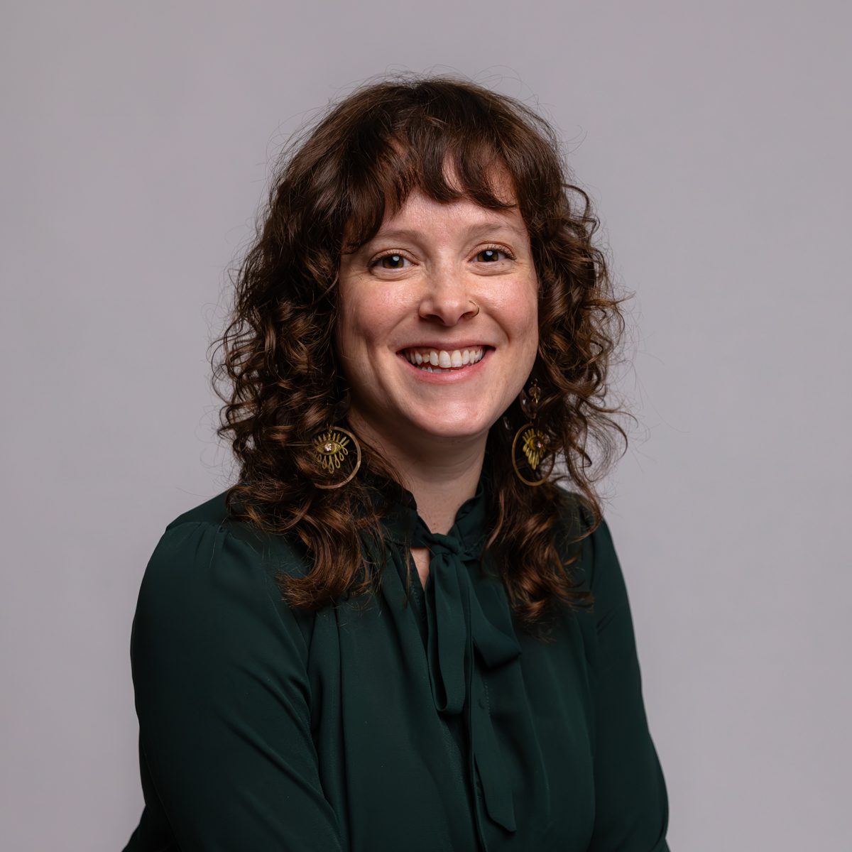 A woman with curly brown hair and bangs, wearing a dark green blouse with a bow and large circular earrings, smiling in front of a plain light gray background.