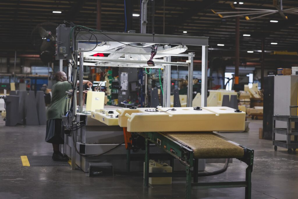 A person operates machinery in a large factory, working with light-colored plastic parts on a conveyor belt. The factory is spacious with high ceilings and industrial lighting.