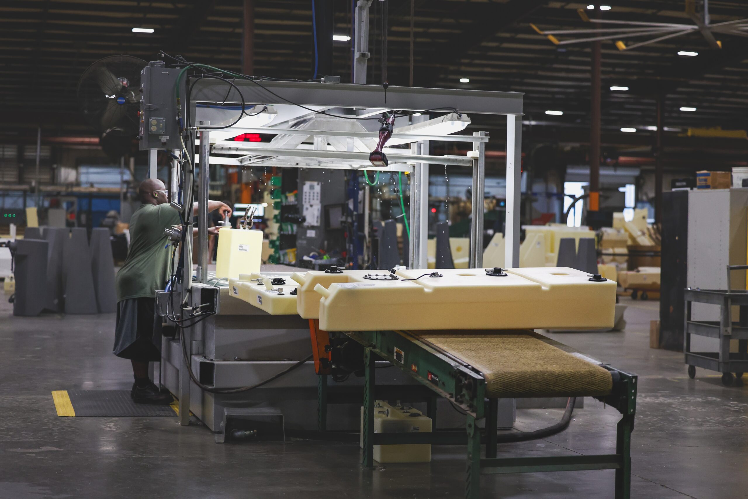A person operates machinery in a large factory, working with light-colored plastic parts on a conveyor belt. The factory is spacious with high ceilings and industrial lighting.