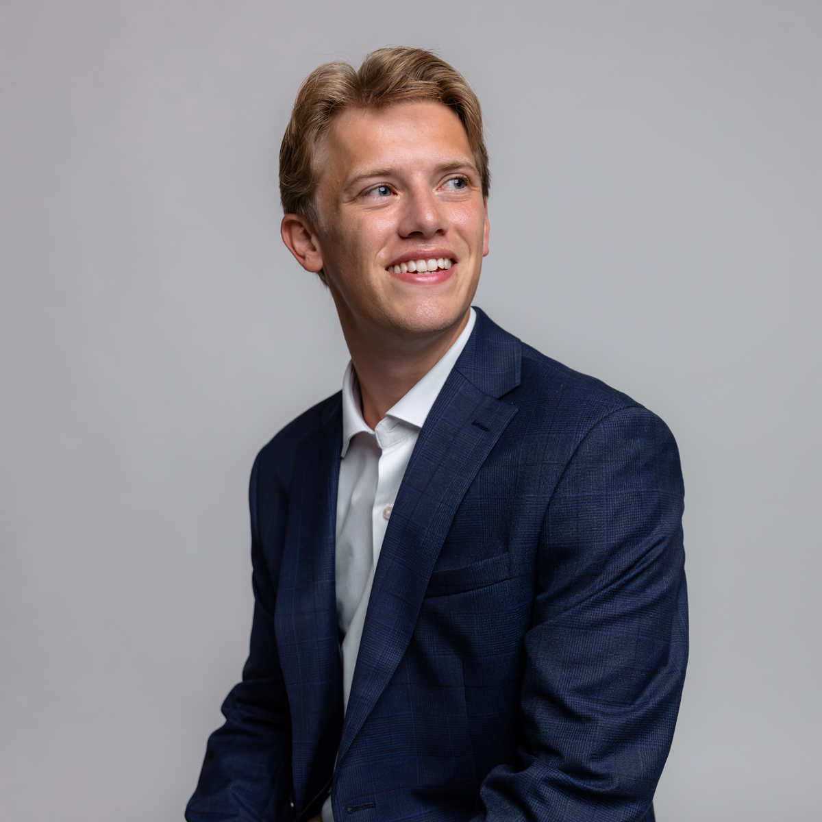 Young man in a navy blue suit and white shirt sits and smiles, looking to his left, against a plain light gray background.