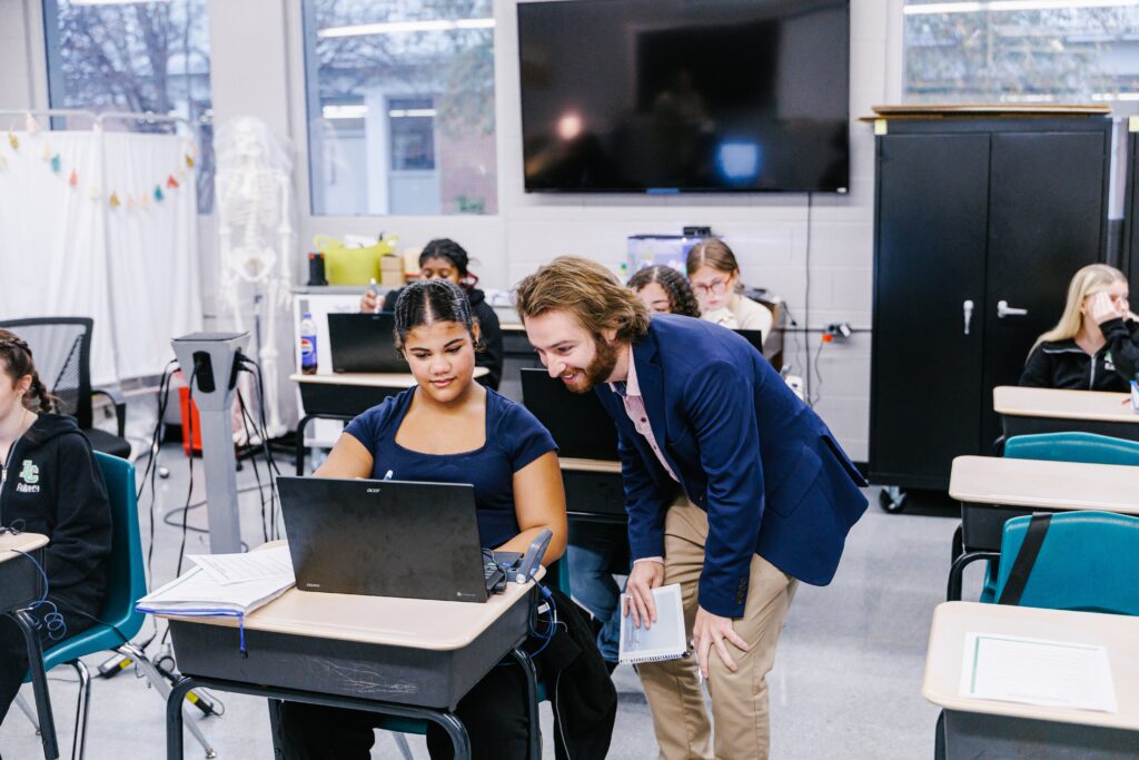 A teacher in a blue blazer leans over to help a student working on a laptop in a classroom. Other students are seated at desks, and a TV and cabinets are visible in the background.