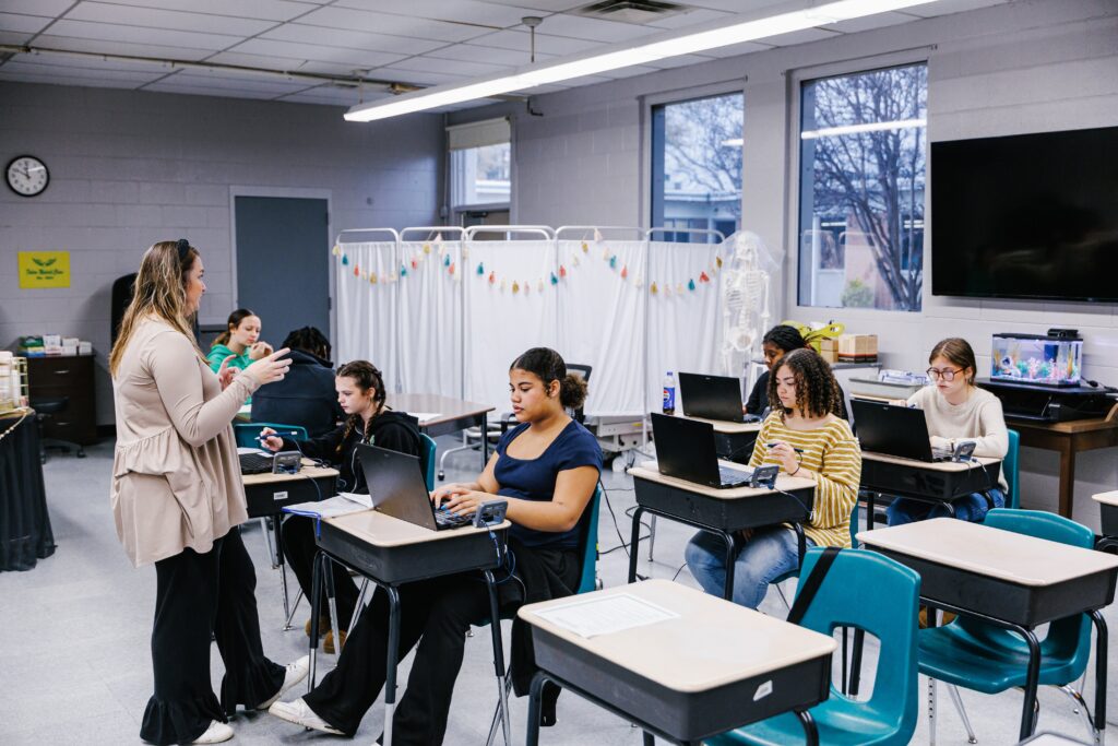 A teacher stands and speaks to students seated at desks using laptops in a classroom with medical mannequins and equipment in the background.