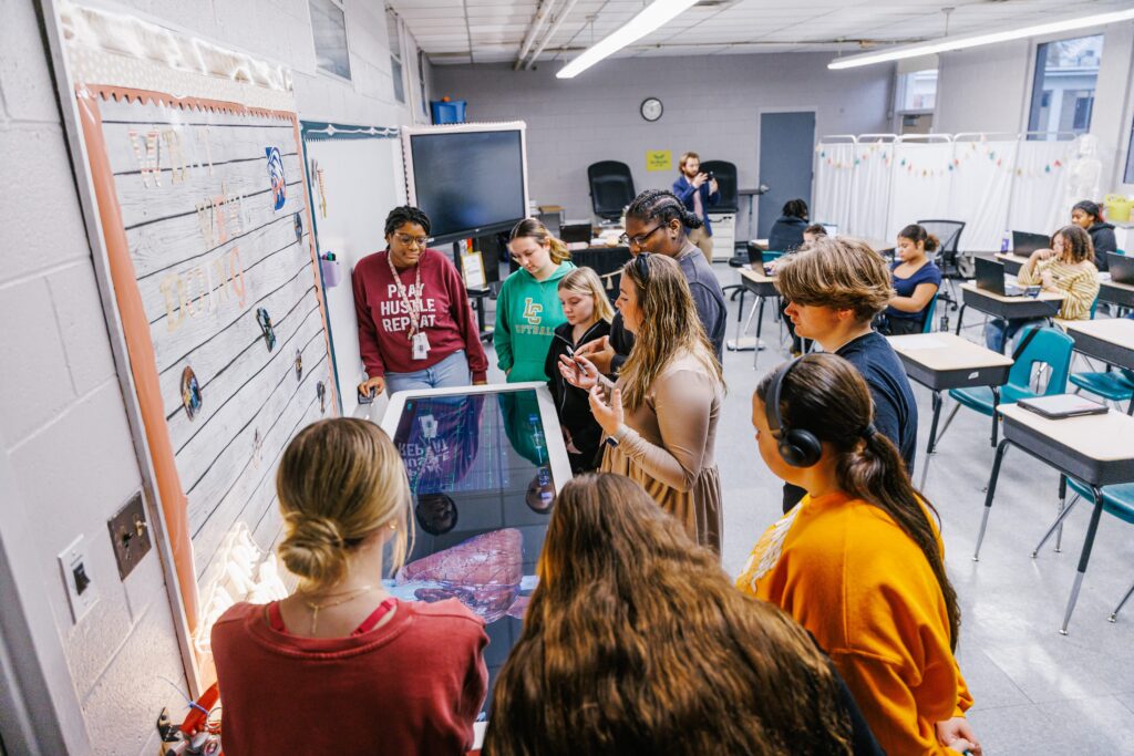 A group of students and a teacher gather around an interactive digital anatomy table in a classroom, observing and discussing a virtual human body display.